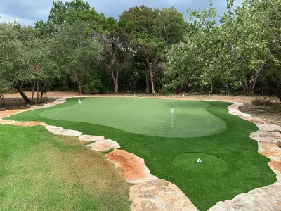 Custom backyard putting green with multiple practice holes, artificial grass fringe, stone edging, and hitting area